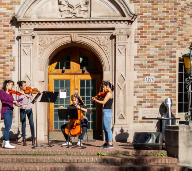 Four students playing stringed instruments in front of a UW building