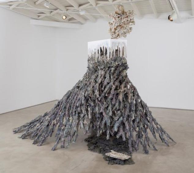 A right-up-triangular-shaped work sits in the foreground of a concrete gray gallery space.